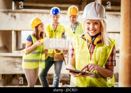 Belle jeune femme architectes construction using digital tablet sur un chantier de construction. Plan d'examen de ses collègues en arrière-plan. Banque D'Images
