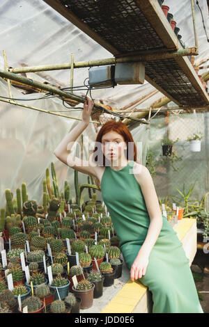 Jeune fille rousse en robe vert assis sur la tablette avec cactus et à sérieusement l'appareil photo Banque D'Images