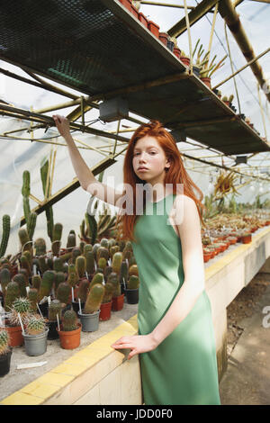 Jeune fille rousse en robe vert assis sur la tablette avec cactus et à sérieusement l'appareil photo Banque D'Images