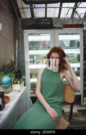 Belle jeune fille rousse en robe verte à regarder caméra dans la verdure contre frigidaires avec plantes Banque D'Images