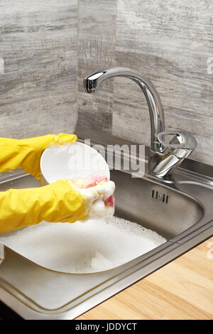 Close up de femmes dans les mains des gants en caoutchouc de protection jaune bol blanc laver avec une éponge de nettoyage rose contre l'évier de cuisine pleine de mousse Banque D'Images