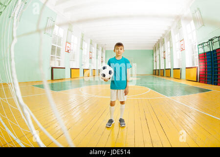 Portrait en pied de 12 ans garçon en uniforme de football, debout à l'école de sport, holding soccer ball, une balle dans le filet de la porte Banque D'Images