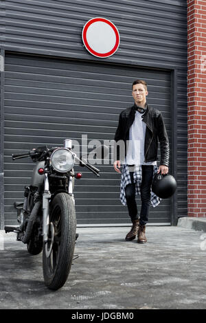 Beau jeune homme en noir veste biker bottes et aller à son style classique cafe racer motorcycle portes industrielles comme arrière-plan. Ma coutume de vélo Banque D'Images