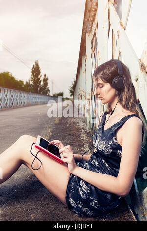 Belle jeune femme à l'écoute de la musique avec tablet et assis sur la route Banque D'Images