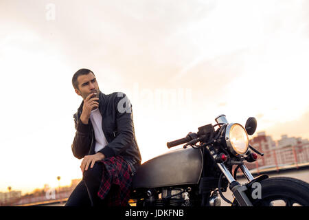 Handsome Guy Rider avec barbe et moustache en noir veste biker fumeurs tandis que s'asseoir sur un style classique du cafe racer moto au coucher du soleil. J'ai fait sur commande de vélo Banque D'Images