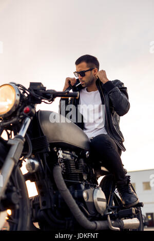 Beau jeune mec avec barbe et moustache en noir lunettes fashion 1900 fumeurs et corriger biker jacket s'asseoir sur un style classique du Cafe Racer Banque D'Images