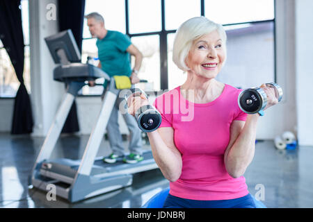 La sportive senior sitting on fitness ball avec haltères, sportif sur tapis roulant derrière dans la haute classe de conditionnement physique Banque D'Images