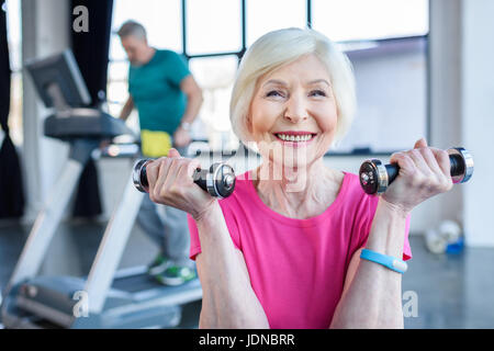 La sportive senior sitting on fitness ball avec haltères, sportif sur tapis roulant derrière dans la haute classe de conditionnement physique Banque D'Images