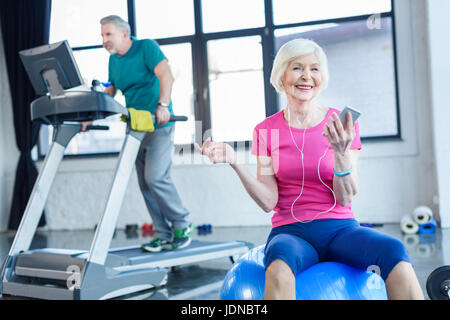 La sportive senior sitting on fitness ball avec le smartphone, sportif sur tapis roulant derrière dans la haute classe de conditionnement physique Banque D'Images