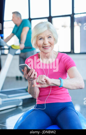 La sportive senior sitting on fitness ball avec le smartphone, sportif sur tapis roulant derrière dans la haute classe de conditionnement physique Banque D'Images