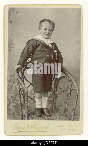 Original CDV (carte de visite) portrait studio de jeune garçon victorien souriant portant un costume de marin tendance debout sur une chaise en osier, toile de fond peinte, Truro, Cornwall, Royaume-Uni vers 1890 Banque D'Images