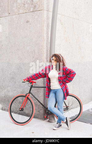 Jeune belle mixed race woman listening music avec des écouteurs s'appuyant sur son vélo - sportive, concept musique Banque D'Images