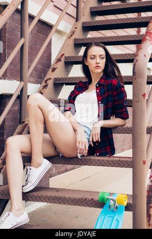 Belle jeune femme en chemise à carreaux et jeans taille basse short shorts et baskets blanches s'asseoir sur l'escalier avec son mini bleu skateboard longboard. Banque D'Images