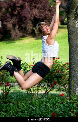 Happy young woman se balançant d'un arbre dans le jardin des coups de talons dans l'air comme elle rit à l'appareil photo Banque D'Images