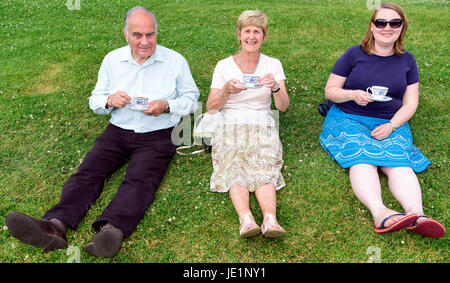 Trois personnes lors d'une fête du village local assis sur l'herbe prendre une tasse de thé sur une chaude journée d'été, Herriard, Hampshire, Royaume-Uni. 18 juin 2017. Banque D'Images