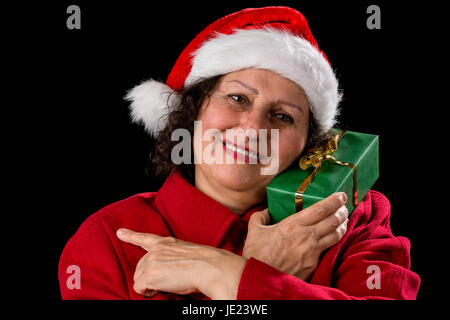 Heureux avec une femme âgée Santa Claus hat et un manteau rouge. Elle est titulaire d'un cadeau de Noël vert enveloppé de sa joue gauche, tout en pointant avec son index gauche sur sa droite. Fond noir. Banque D'Images