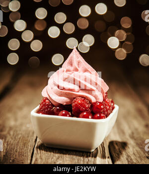 Les framboises et groseilles servi dans un plat garni d'un tourbillon de yaourt petits fruits congelés sur un comptoir de bar en bois contre un bokeh de lumières scintillantes lumières parti Banque D'Images