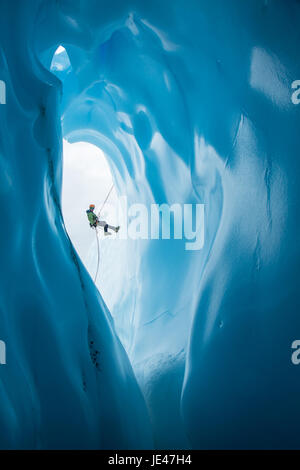 Un grimpeur sur glace dans une gaine verte et orange helmet rappels passé une grande entrée arrondie à une grotte de glace sur le Glacier Matanuska en Alaska. Banque D'Images