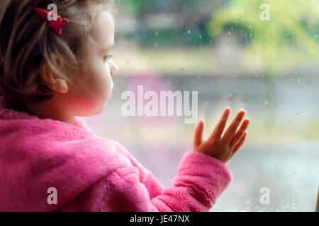 Gros plan d'une petite fille mignonne en rose peignoir regarde tristement par la fenêtre. Assis sur le rebord de la fenêtre. Banque D'Images