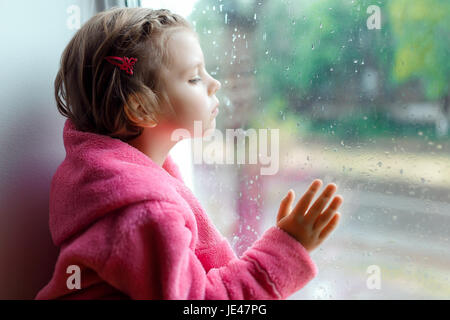 Gros plan d'une petite fille mignonne en rose peignoir regarde tristement par la fenêtre. Assis sur le rebord de la fenêtre. Banque D'Images