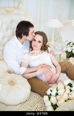 Beau couple, jeune femme enceinte et l'homme serrant avec amour baisers, assis sur le lit, dans une maison de l'intérieur. Banque D'Images