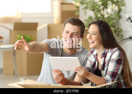 Couple heureux déménagement maison ensemble sur la ligne de planification avec une tablette assis sur le plancher à la maison avec des boîtes à l'arrière-plan Banque D'Images