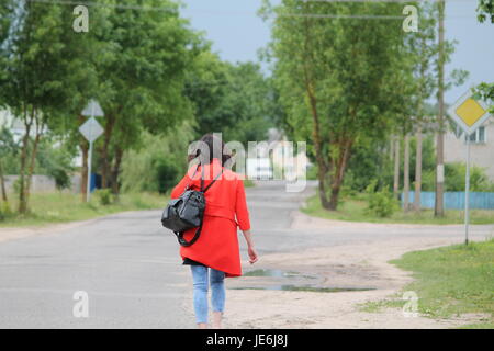 Jeune femme cheveux sombres en manteau rouge marche rapide dans l'allée d'été vert Banque D'Images