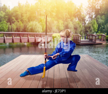 Wushu fighter féminin avec lame contre lake et vert nature, arts martiaux femme en tissu bleu sur l'entraînement au combat en plein air Banque D'Images