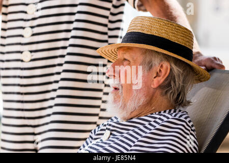 Vue de côté de man, 60, avec barbe grise assis dans un transat à l'ombre. Tête et épaules. Le port d'un chapeau de paille et plage de style Victorien costume. Banque D'Images