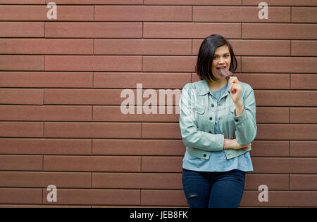 Jeune et jolie brunette woman dans une veste en cuir gris goûte la crème glacée en lustre de chocolat près d'un mur de brique brune texturée. Banque D'Images