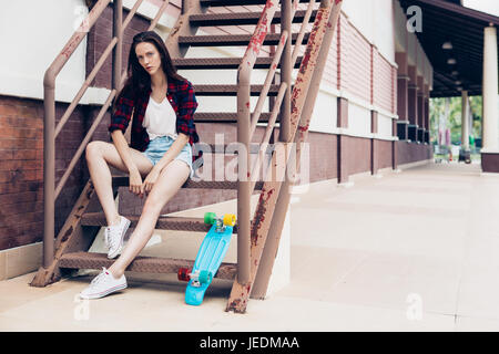 Belle jeune fille en chemise à carreaux et jeans taille basse short shorts et baskets blanches s'asseoir sur l'escalier avec son mini bleu skateboard longboard. U Banque D'Images