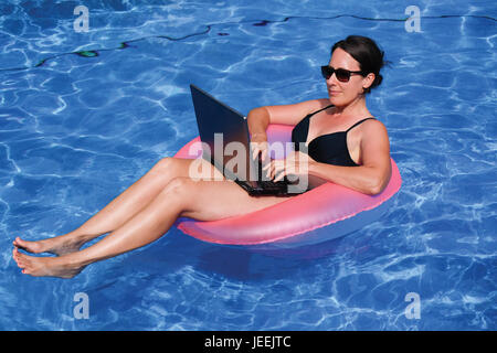 Les femmes d'âge moyen de la bourreau de travail sur ordinateur portable en rose anneau de sauvetage gonflable dans une piscine en vacances Banque D'Images