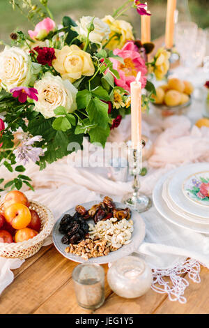 Aire de pique-nique, de l'alimentation, l'été, l'appartement de concept - close up sur la table à la décoration de fête avec beau bouquet de roses et de pivoines rose, des bougies dans les chandeliers, les fruits secs, les nectarines, les noix, la verrerie. Banque D'Images