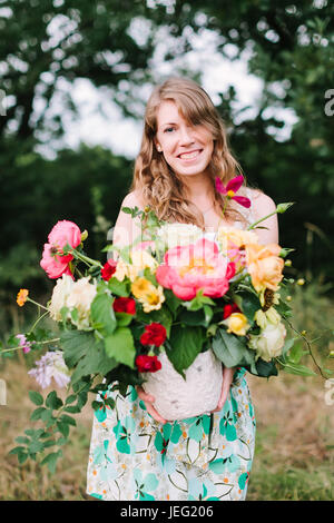 Mariage, célébration, la beauté, la vie à la campagne, le printemps, la nature concept - Jeune femme séduisante avec les cheveux brun clair dans cette robe à imprimé floral puting out joli bouquet de fleurs divers Banque D'Images