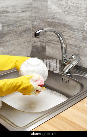 Close up de femmes dans les mains des gants en caoutchouc de protection jaune bol blanc laver avec une éponge de nettoyage rose contre l'évier de cuisine pleine de mousse Banque D'Images