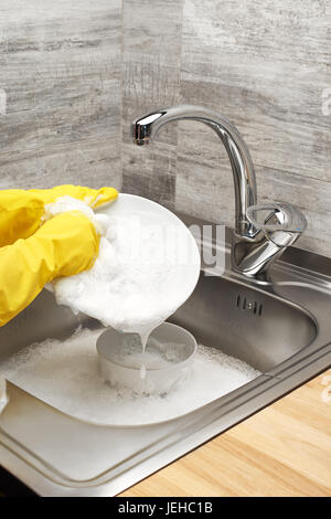 Close up de femmes dans les mains des gants en caoutchouc de protection jaune lave-plaque blanche contre l'évier de cuisine pleine de mousse et de la table Banque D'Images