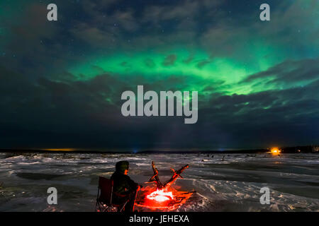 Un homme se réchauffe lui-même à côté d'un feu de bois flotté tout en regardant l'aurore à travers les nuages sur la rivière Delta, près de Delta Junction, Alaska. Banque D'Images