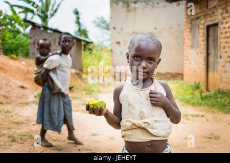 Un jeune enfant est maintenant un morceau de fruit avec une fille tenant un jeune enfant sur le dos en arrière-plan ; l'Ouganda Banque D'Images