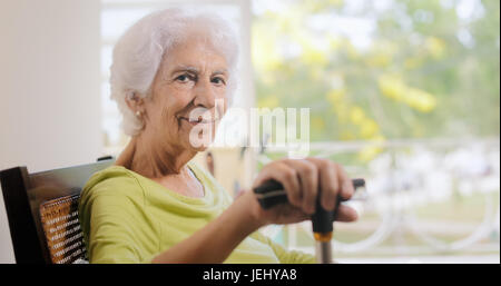 Portrait de vieille dame assise sur la bascule à la maison. Happy elderly woman relaxing on rocking chair et la tenue de bâton de marche. Copy space Banque D'Images