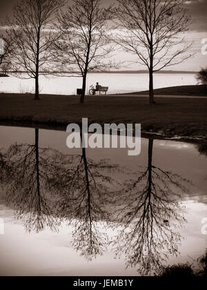 Pour le coucher du soleil monochrome : Un homme est assis sur un banc et bénéficie d'un printemps chaud en soirée. Les feuilles ne sont pas encore sur les arbres. Banque D'Images