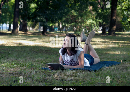 Belle jeune femme allongé dans l'herbe et détend tout en discutant avec vos amis via tablet Banque D'Images