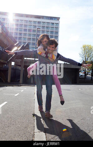 Jeune couple s'amusant dans la ville. La femme est sur son dos et les deux partenaires ont leurs bras tendus tout en regardant la caméra. Banque D'Images