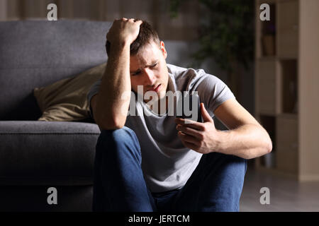 Seul homme triste checking mobile phone assis sur le plancher dans la salle de séjour à la maison avec un fond sombre Banque D'Images