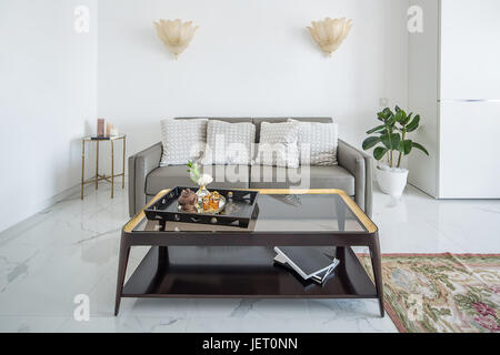 Intérieur moderne blanc avec carrelage au sol avec un tapis. Il y a des lampes dorées fantaisie sur le mur gris, canapé avec coussins, table avec une table en verre wi Banque D'Images