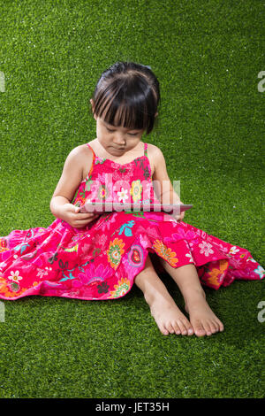 Chinois asiatique petite fille assise sur l'herbe et jouer au parc de plein air de l'ordinateur tablette Banque D'Images