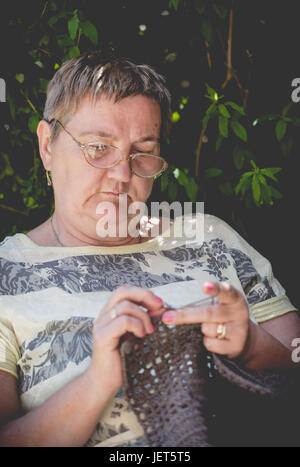 Senior woman, cheveux courts, le crochet d'une couverture marron se détendre dans le jardin. Composition verticale se concentrer sur visage concentré Banque D'Images