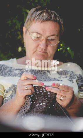 Senior woman, cheveux courts, le crochet d'une couverture marron se détendre dans le jardin. Composition verticale, se fonder sur les mains Banque D'Images