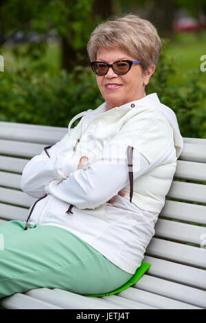 Smiling femme européenne dans l'âge assis sur un banc de parc, les bras croisés sur la poitrine Banque D'Images