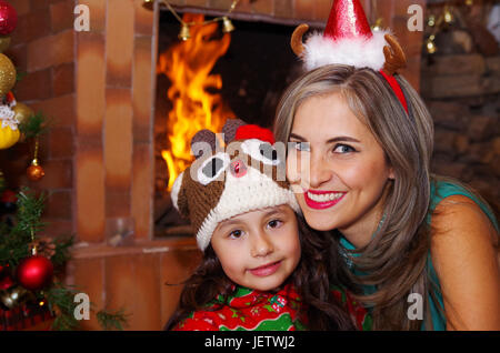 Mère et fille heureux à Noël, litle girl wearing a deer hat et maman un chapeau de Noël, avec une chimmey arrière-plan. Banque D'Images