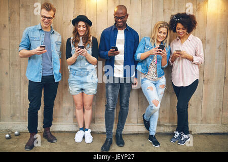 Cheerful group of friends standing en noir et blanc et de la navigation sur leurs smartphones ensemble au mur en bois. Banque D'Images